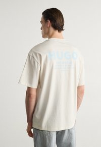 Beige, nagy méretű pamut póló, világoskék "HUGO" nyomtatott logóval és szöveggel a hátán, rövid ujjal és kerek nyakkivágással.
