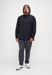 Camicia nera a maniche lunghe con bottoni e tasca sul petto, abbinata a pantaloni cargo grigi e sneakers bianche con accenti neri.