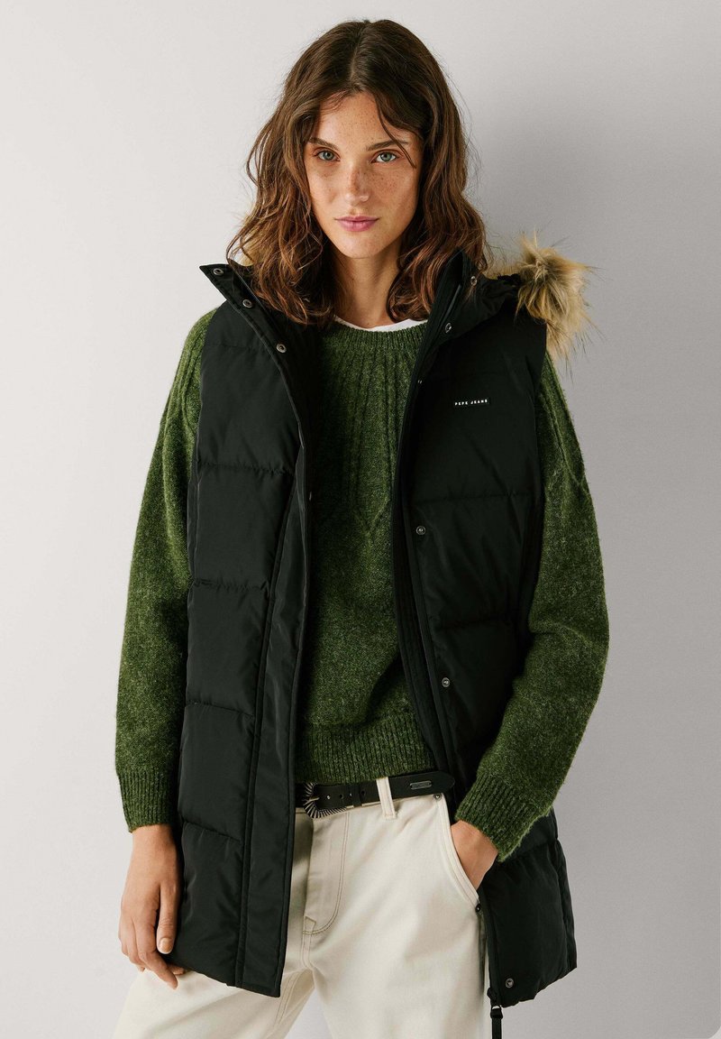 Melna piepūsta veste ar kažokādas odere kapuci, uzvilkta virs zaļas adītas džempera. Aprīkot ar pogām un sānu kabatām, savienota ar bežām biksēm.