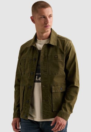 Homme portant une veste boutonnée vert olive sur un t-shirt graphique beige, regardant légèrement sur le côté sur un fond uni.