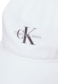 Casquette en coton blanc avec une visière cousue. L'avant présente un logo "CK" noir avec le texte "Calvin Klein Jeans" en gris.