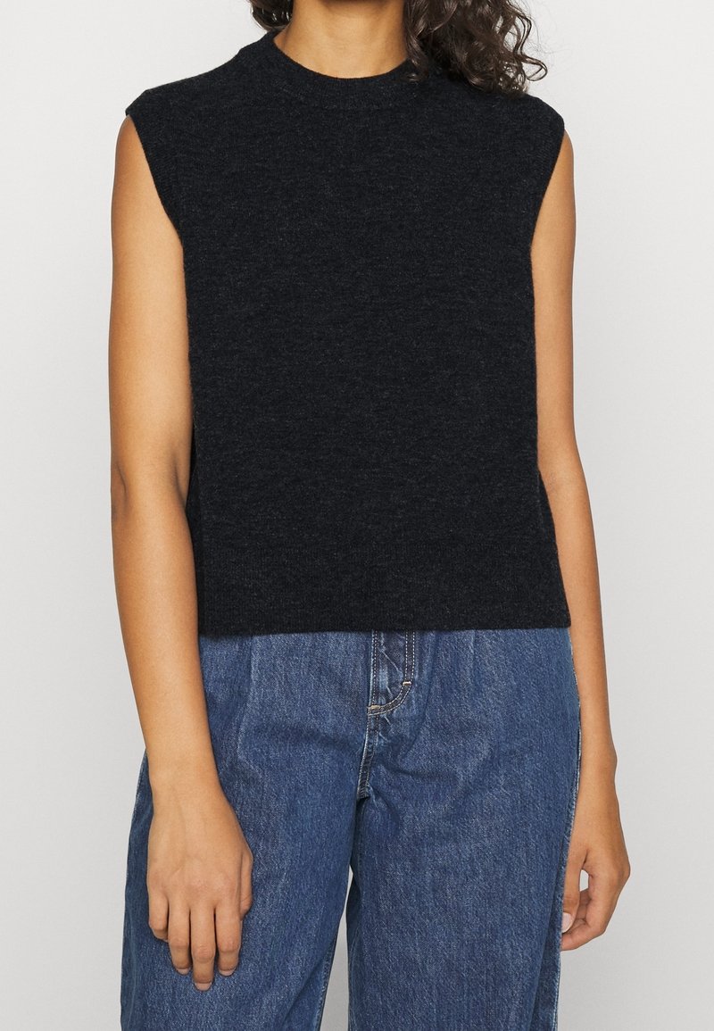 Persona che indossa un top senza maniche nero a maglia con un collo alto e jeans blu a vita alta, contro uno sfondo semplice.