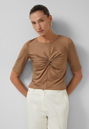 Frau mit nach hinten gegelten Haaren, die goldene Ohrringe, ein braunes, vorne gedrehtes Kurzarmtop und hoch taillierte weiße Hosen trägt, steht vor einer grauen Wand.