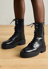 Botas de cuero negro con cordones y suelas gruesas, cremallera lateral y ojetes de metal. Acabado suave con textura variada en el caño.