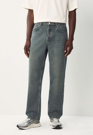 Mann trägt ausgewaschene blaue Jeans mit geradem Bein, ein weißes Kurzarmhemd und graue-weiße Sneakers, steht vor einem schlichten Hintergrund.