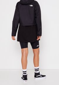 Personne debout, de dos, portant une veste noire cropped, un short de vélo noir, des chaussettes blanches avec des rayures noires et des baskets noires montantes.