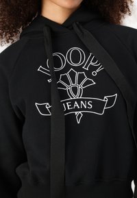 Black hoodie featuring white embroidery with the text "LOOP! JEANS" and a decorative design. Includes a drawstring and ribbed hem.