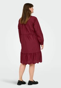 Burgundy cotton dress with long sleeves, gathered waist, and scalloped eyelet hem. Model wearing black shoes, viewed from the back.