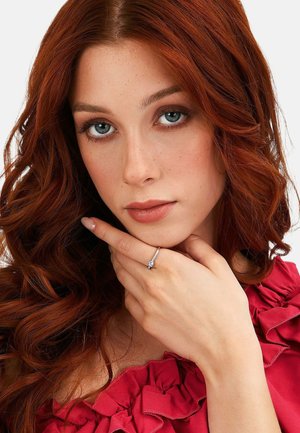 Junge Frau mit roten lockigen Haaren und blauen Augen, die ein rotes Rüschenoberteil trägt und einen silbernen Ring mit Diamant an ihrem Finger nahe ihrem Gesicht zeigt.