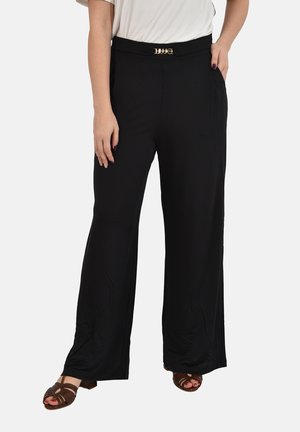 Donna che indossa pantaloni neri a gamba larga con dettaglio fibbia dorata abbinati a top bianco e sandali marroni open-toe, in piedi su sfondo bianco.