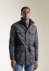 Parka de color azul oscuro con un cuello alto, cierres de cremallera y a presión, cuatro bolsillos frontales y una tela texturizada. Llevada sobre una camisa con cuello.