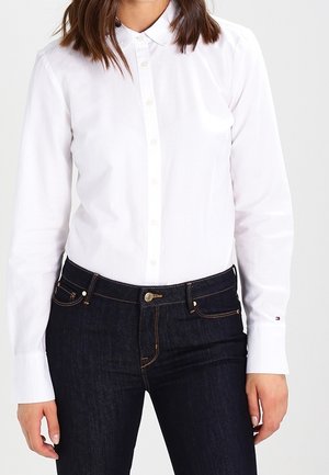 Femme portant une chemise blanche ajustée avec un petit col, associée à un jean taille haute bleu foncé avec des coutures dorées.