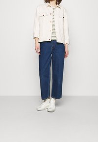 Lys beige denimjakke med to brystlommer, parret med en stribet skjorte i oliven og hvid, mørkeblå brede jeans og hvide sneakers.