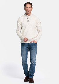 Man in een beige knooptrui, blauwe spijkerbroek en bruine suède laarzen, staand met één hand in de zak tegen een witte achtergrond.
