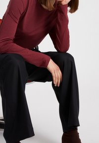 Bordeaux long-sleeve shirt, gecombineerd met losse zwarte broek. Soepele stofstructuur, minimalistisch ontwerp. Hand rustend op de knie.