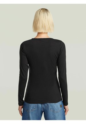 Personne aux cheveux blonds courts portant un haut noir à manches longues et un jean bleu, debout de dos sur un fond clair uni.