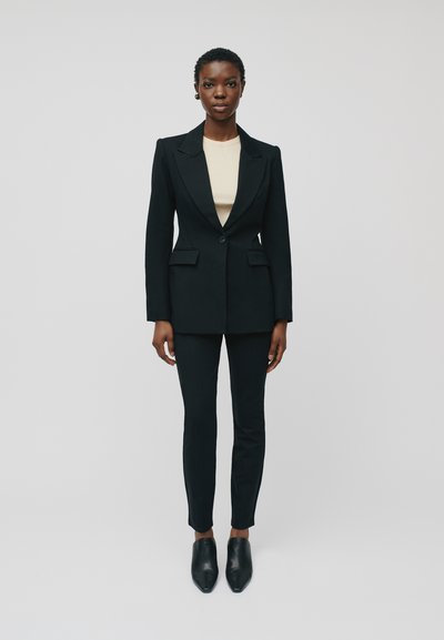 Sort skræddersyet blazer med slåede revers, parret med matchende smalle bukser. Underneath en beige top. Afsluttet med sorte kantede sko.