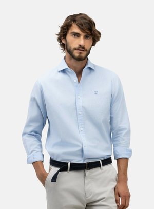 Hombre con cabello corto y castaño y barba, que lleva una camisa azul claro con botones y pantalones beige con cinturón negro, está de pie frente a un fondo liso.