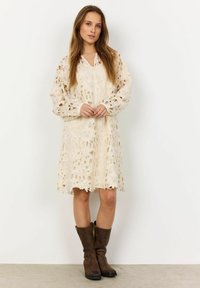 Krajkov dress v off-white farbe s kvetinovými výrezmi, dlhými rukávmi a golierom; spárované s hnedými vysokými čižmami po kolená. Detail s vlnkovaným okrajom.