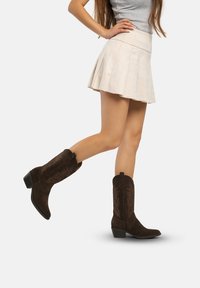 Botas de cowboy de ante marrón con punta afilada y tacón bajo, que presentan patrones bordados detallados a lo largo de la caña.