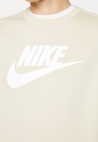 Gros plan d'une personne portant un sweat-shirt beige avec un grand logo Nike blanc et un swoosh sur la poitrine.