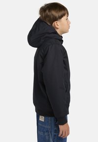 Sort hættejakke med en glat tekstur, elastiske manchetter og sidelommer, iført med blå denimjeans. Sidevisning præsenteret.