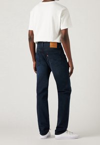Dark blue denim jeans with a straight-leg fit, paired with a white t-shirt, featuring a tan leather patch on the back waistband.