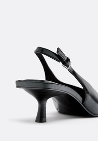 Musta nahkainen slingback-kengä matalalla kitten-korolla ja säädettävällä solkiremillä, esitetty vaaleaa taustaa vasten.