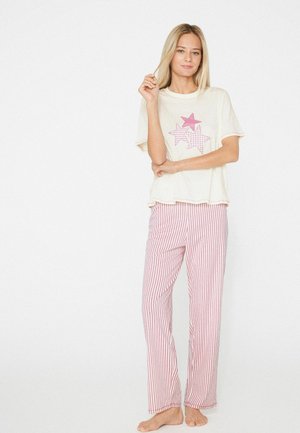 Jonge vrouw staat blootsvoets, draagt een crèmekleurig T-shirt met roze sterren en een roze-wit gestreepte pyjamabroek op een witte achtergrond.