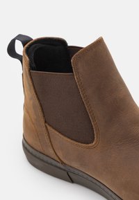 Brown leather ankle boot with elastic side panels, textured finish, pull tab, and a black rubber sole. Close-up view showing details.