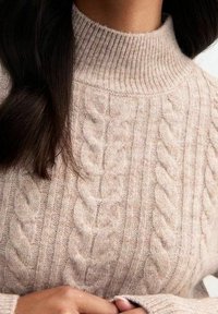 Béžový svetr s rolákem a pleteným vzorem, na kterém je vidět texturu pleteniny a část dlouhých tmavých vlasů.