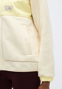 Μπουφάν από φλις σε κρεμ χρώμα με κίτρινο νάιλον επάνω μέρος, ριγέ τελείωμα και πλαϊνή τσέπη με κούμπωμα με κλιπ.