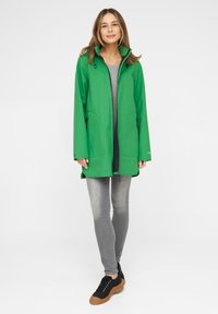 Abrigo impermeable verde con un alto cuello, cierre de cremallera frontal y dos bolsillos laterales, combinado con jeans grises y zapatillas negras. Textura suave.