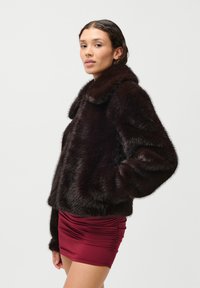 Καφέ ψεύτικη γούνα με στρογγυλό γιακά και φαρδιά μανίκια, συνδυασμένη με μια εφαρμοστή μπορντό μίνι φούστα με πτυχώσεις.