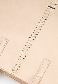 Beige leren handtas met een centrale rij van metallic gouden studs, platte handvatten en nauwkeurige stiksels langs de randen.