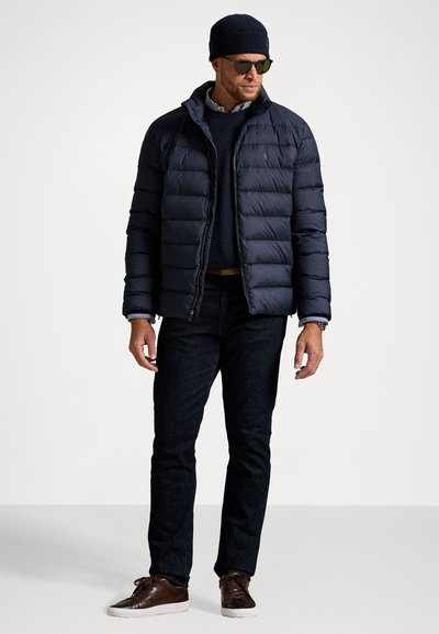 Mand iført en marineblå puffjakke, mørke jeans, brune sneakers, stribet skjorte, marineblå sweater, hue og solbriller, der står mod en hvid baggrund.