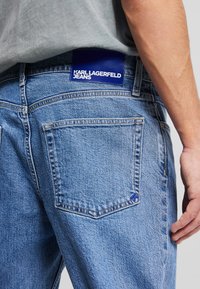 Jean en denim bleu délavé moyen avec une patch en cuir bleu distinctif portant le logo "KARL LAGERFELD JEANS" sur la taille arrière.