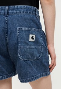 Οπίσθια όψη μπλε τζιν σορτς με τσέπη με κέντημα στο πίσω μέρος που φέρει το λογότυπο Carhartt σε μαύρο και άσπρο, φορεμένα από άτομο με μαύρο τοπ.
