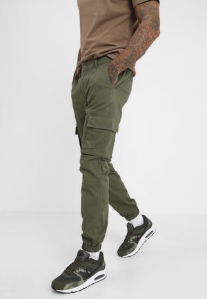 Mężczyzna w oliwkowych joggerach typu cargo, brązowym t-shircie i zielonych sneakersach idzie z rękami w kieszeniach na jednolitym tle.