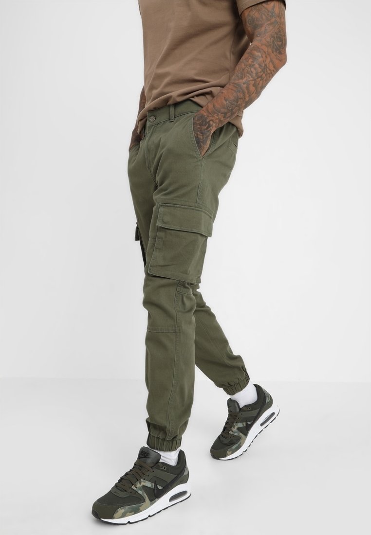 Homme portant un pantalon cargo jogger olive, un t-shirt marron et des baskets vertes, marchant les mains dans les poches devant un fond uni.