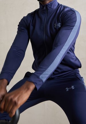 Homme en survêtement bleu marine avec des rayures bleu clair tenant une kettlebell, réalisant un entraînement à l’intérieur.