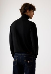 Maglione nero a collo alto con una texture liscia, abbinato a jeans in denim scuri con cuciture gialle e un logo triangolare sulla tasca posteriore.
