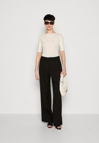 Cremfarbene, figurbetonte Bluse mit Ellenbogenärmeln, kombiniert mit hoch taillierten, schwarzen, weiten Hosen. Die Accessoires umfassen eine weiße Handtasche und schwarze Sonnenbrillen.