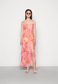 Vestido sem mangas em tons de rosa e laranja, com uma racha alta na coxa, feito de um tecido leve. Combinado com sandálias rosas e uma bolsa pequena.