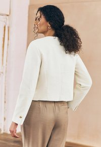 Giacca corta bianca a costine con maniche lunghe, scollatura tonda e dettagli in bottone dorato. Abbinata a pantaloni beige chiaro.
