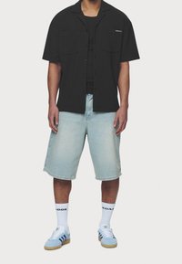 Camicia nera a maniche corte con bottoni e due tasche frontali, abbinata a pantaloncini di denim azzurro chiaro e sneakers azzurro chiaro con dettagli bianchi.