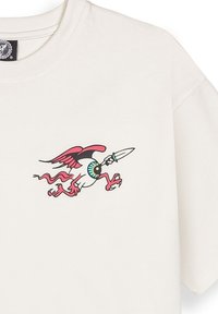 Camiseta de algodón color crema con un gráfico colorido de una criatura alada con un ojo y un cuchillo, situada en el frontal.