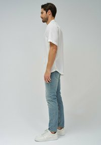 Camicia bianca a maniche corte con colletto, abbinata a jeans slim-fit azzurri chiari e sneaker bianche, mostrata da un profilo laterale.