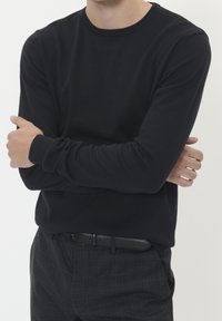 Mies, joka käyttää mustaa pitkähihasta villapaitaa ja tummia ruudullisia housuja mustalla vyöllä, kädet ristissä rinnalla, tasaisella taustalla.