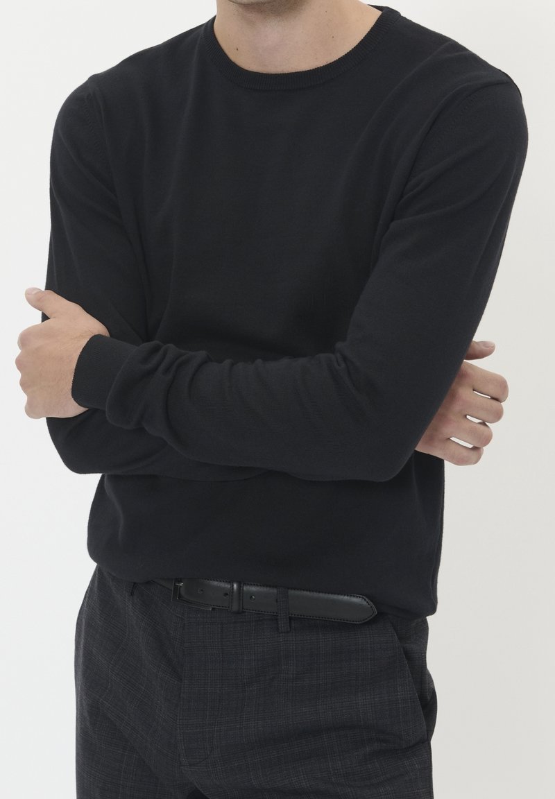 Mies, joka käyttää mustaa pitkähihasta villapaitaa ja tummia ruudullisia housuja mustalla vyöllä, kädet ristissä rinnalla, tasaisella taustalla.